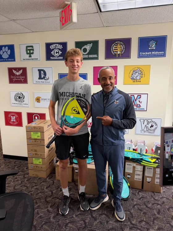 Alex Forest presenting a new tennis racquet to a Detroit Public Schools athletics staff member alongside cases of donated racquets and balls